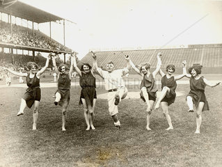 Mann und sechs Frauen tanzen auf Rasen eines Stadions
