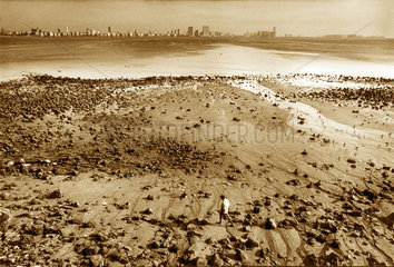 Indien - Mann an steinigem Strand von Bombay mit Skyline im Hintergrund