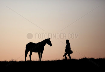 Gestuet Goerlsdorf  Silhouette  Mann geht bei Morgendaemmerung auf einer Weide auf ein Pferd zu