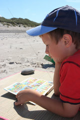 Hvide Sande  Daenemark  Junge mit Schirmmuetze liest am Strand