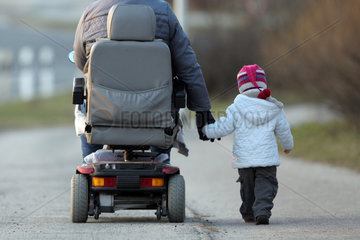 Handewitt  Deutschland  eine Grossmutter faehrt in einem elektrischen Rollstuhl mit ihrem Enkel an der Hand auf einem Fahrradweg spazieren