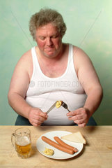 Dicker Mann in Unterhemd isst zwei Wuerstchen mit Senf