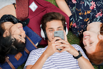 Young man lying on grass with friends  using smartphone and listening to headphones
