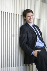 Businessman leaning against wall with hands in pockets  smiling cheerfully