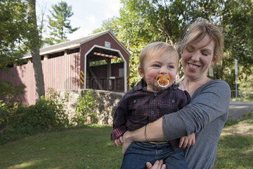 Mother holding toddler son in front of covered bridge  portrait