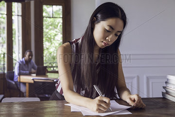 Young woman writing on paper while man watches her in the background