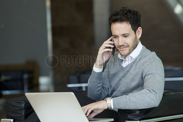 Businessman using cell phone and laptop computer at hotel lobby