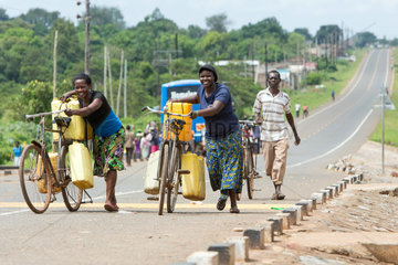 Kamdini  Uganda - Stadtansicht. Zwei Frauen schieben  mit Plastikkanistern beladene Fahrraeder einen Huegel hinauf.