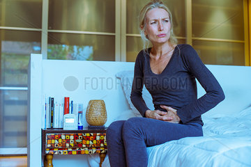 Mature woman sitting on bed with hands on stomach and pained expression on face