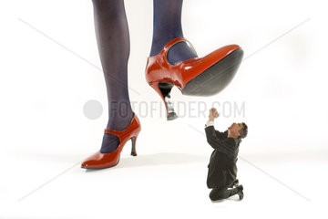 Frau in roten Stoeckelschuhen tritt auf winzigen Mann der um Gnade fleht