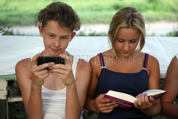 Bolsena  Italien  Junge spielt mit seinem Smartphone waehrend ein Maedchen in einem Buch liest