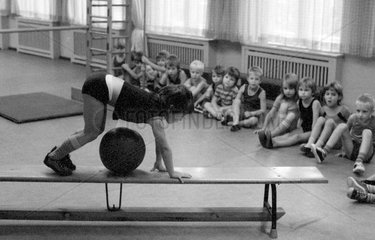 Berlin  DDR  kleines Maedchen klettert im Sportunterricht auf einer Bank ueber einen Medizinball
