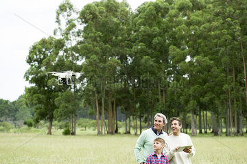 Man flying remote control drone in open field while older man and boy watch