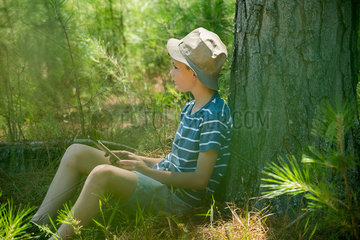 Boy leaning against tree trunk with digital tablet in hands