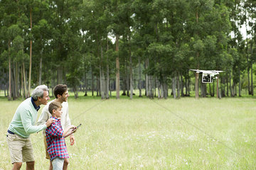 Boy flying remote control drone in open field while father and grandfather watch