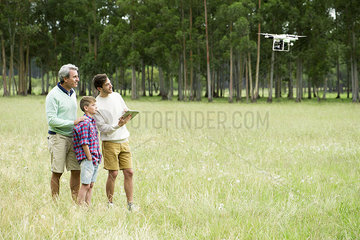 Man flying remote control drone in open field while older man and boy watch