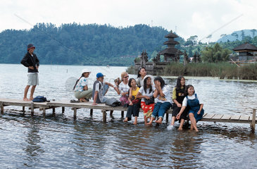 Jugendliche Touristen sitzen auf einem Steg vor dem Tempel Pura Ulun Danu Bratan