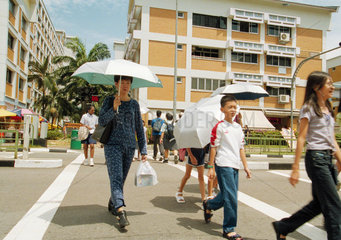 Fussgaenger mit Sonnenschirmen ueberqueren eine Strasse in Singapur