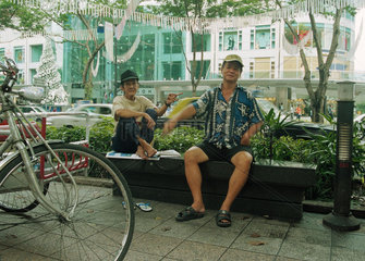 Zwei Maenner sitzen auf einer Bank am Rande der Haupteinkaufsstrasse in Singapur