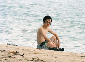 Junger Mann sitzt am Strand eines Erholungsparks in Singapur