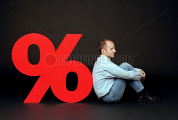Mann sitzt hockend neben einem grossen Prozentzeichen
