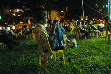 Zuschauer sitzen nachts vor der Buehne eines Strassentheaters in Singapur