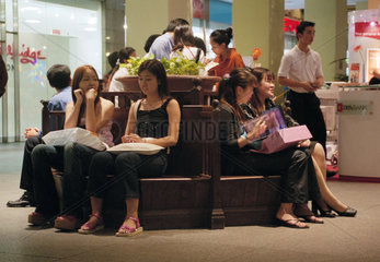 Junge Frauen sitzen abends auf einer Bank in einer Einkaufspassage in Singapur