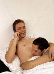 Ein homosexuelles Paar liegt gemeinsam im Bett  einer telefoniert