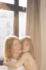 Mother and young daughter embracing by window