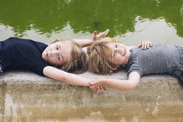 Little girls lying together beside pond  holding hands
