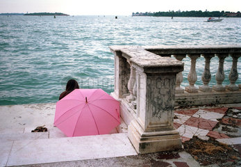 Ein Maedchen sitzt unter einem Regenschirm am Wasser