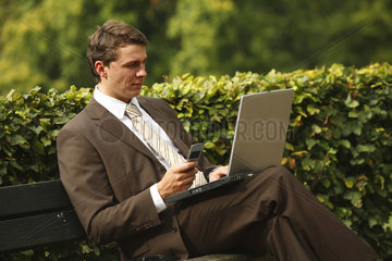 Deutschland  ein junger Mann sitzt im Park an seinem Laptop und telefoniert