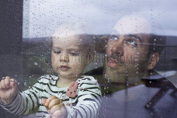 Man with young child looking through window watching rain fall