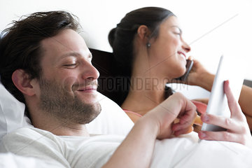 Couple relaxing in bed  man using digital tablet while woman makes phone call
