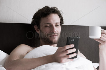 Man in bed using smartphone and drinking hot drink