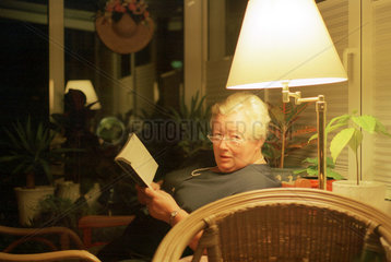 Aeltere Frau sitzt in ihrem Wintergarten und liest ein Buch
