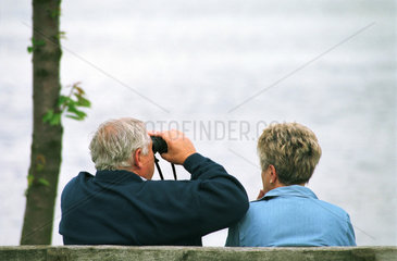 Rentnerpaar sitzt am Wasser mit Fernglas