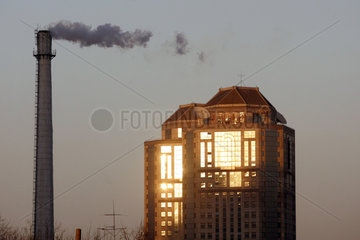 Symbolfoto  qualmender Fabrikschornstein mit einem in der Sonne leuchtenden Wohnhaus
