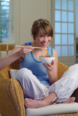 Junge Frau isst zu Hause mit Staebchen ein asiatisches Reisgericht aus einer Schale