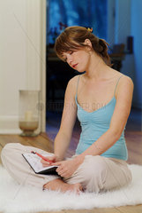 Junge Frau sitzt schreibend zu Hause auf einem Flokati