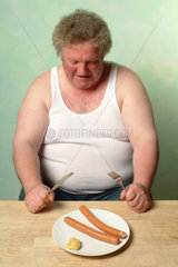 Dicker Mann in Unterhemd sitzt vor einem Teller mit zwei Wuerstchen und Senf