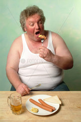 Dicker Mann in Unterhemd isst von einem Teller mit zwei Wuerstchen  Senf und Bier
