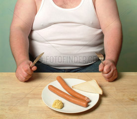 Dicker Mann in Unterhemd sitzt mit zwei Wuerstchen und Senf