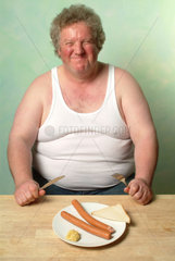 Dicker Mann in Unterhemd sitzt vor einem Teller mit zwei Wuerstchen und Senf