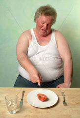 Dicker Mann in Unterhemd sitzt vor einem Teller mit einer halben Tomate