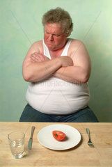 Dicker Mann in Unterhemd sitzt vor einem Teller mit einer halben Tomate