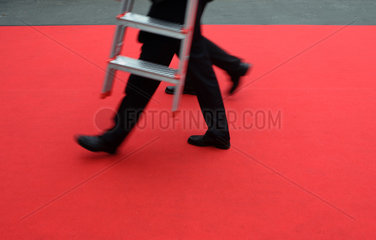 Geschaeftsleute gehen mit einer Leiter ueber einen roten Teppich