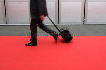 Ein Geschaeftsmann geht mit einem Trolley ueber einen roten Teppich