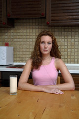 Junge Frau sitzt in ihrer Kueche am Tisch bei einem Glas Milch