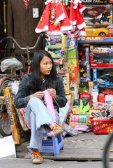 Eine Verkaeferin sitzt vor einem Spielzeuggeschaeft in der Altstadt von Hanoi